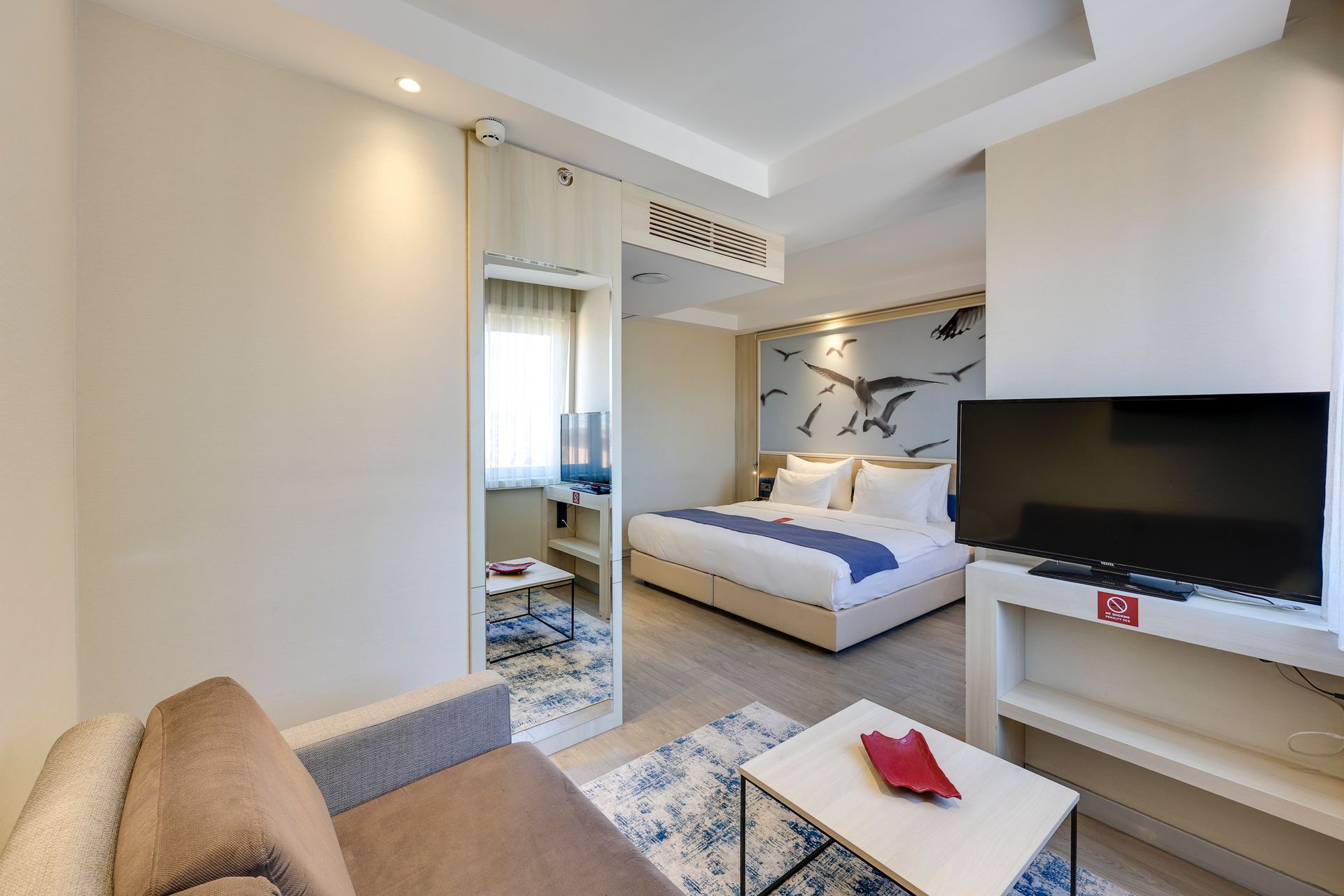 Hotel room with bed, couch, and TV. Neutral colors, modern design, and a bird mural.