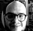 Um homem de óculos e barba está sorrindo em uma foto em preto e branco.