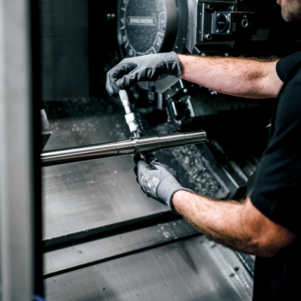 Person mit Handschuhen arbeitet in einer Produktionsumgebung mit einer Maschine an einer Metallstange.