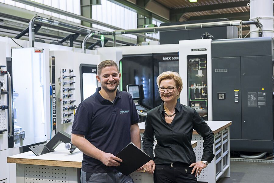 Ein Mann und eine Frau lächeln in einer Fabrik. Sie stehen neben Industriemaschinen.