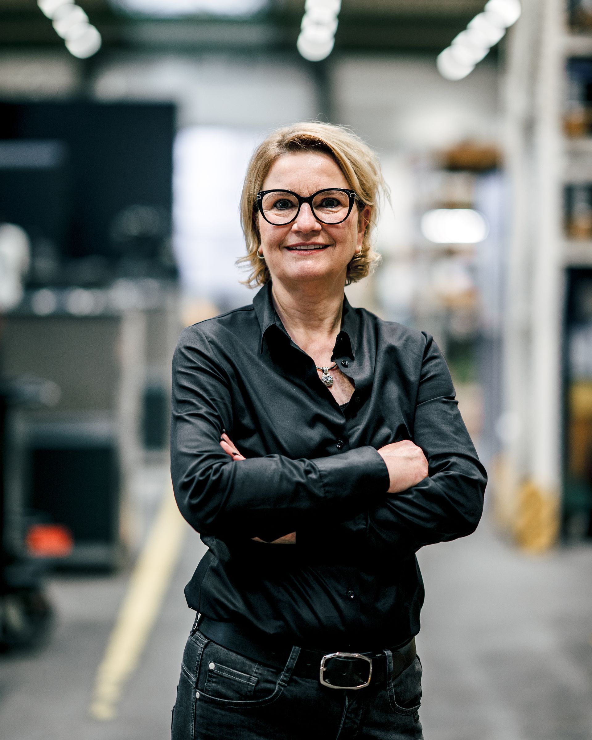 Frau in schwarzem Hemd und Brille mit verschränkten Armen in einer Lagerhalle.