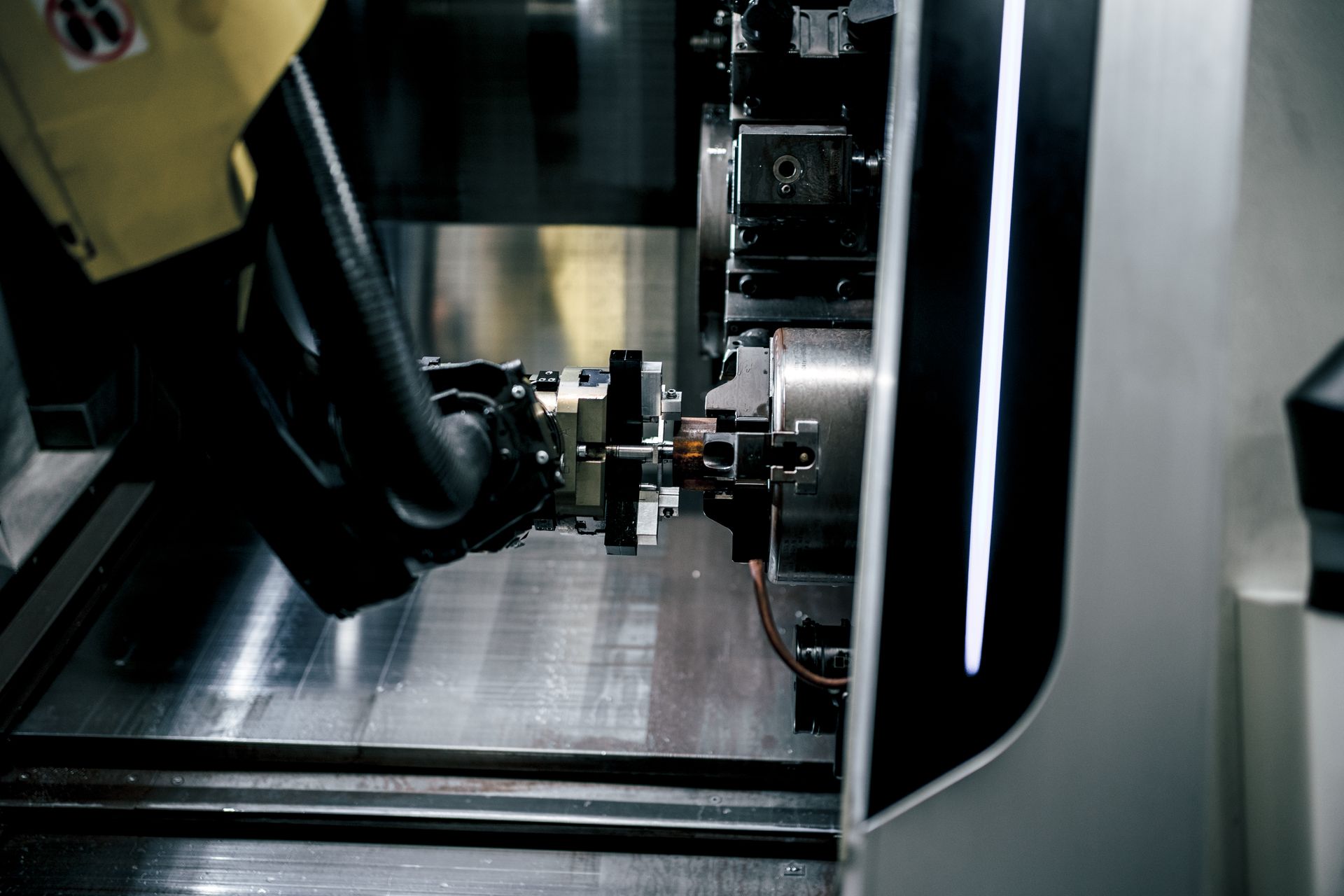Metalldrehmaschine bearbeitet ein kleines Teil, Wasser wird versprüht, im Hintergrund ist der Arm einer Person mit gelbem Ärmel zu sehen.