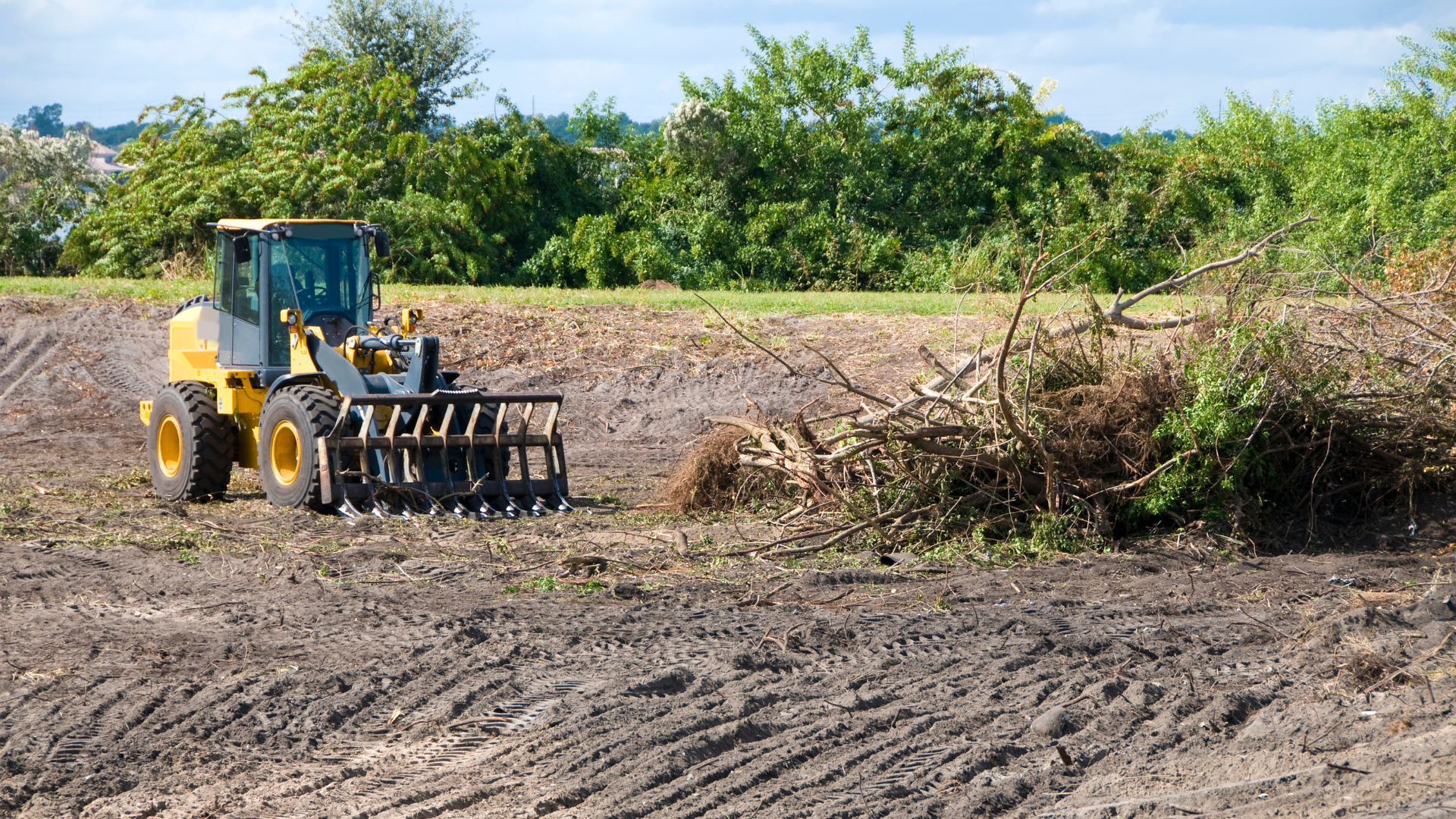 Yellow bulldozer clearing dirt in a muddy field beside a pile of brush and trees