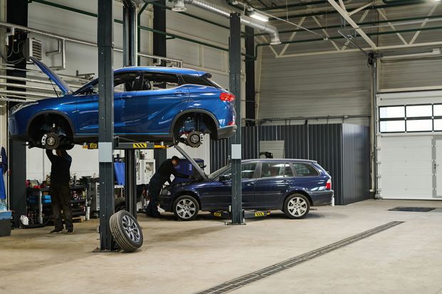 Blue SUV on a lift in an auto repair garage, with another dark car parked below. Blue SUV on a lift in an auto repair garage, with another dark car parked below.