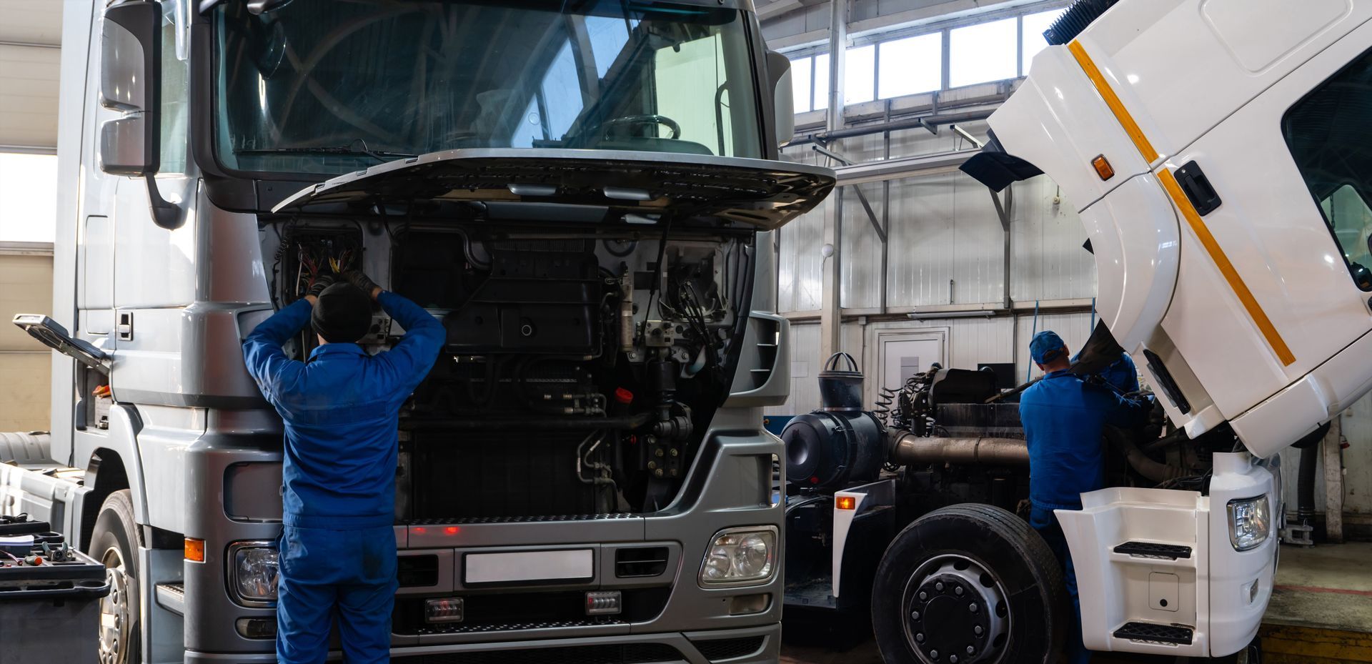 Two mechanics in blue coveralls inspect white truck cabs in an industrial garage. Two mechanics in blue coveralls inspect white truck cabs in an industrial garage.