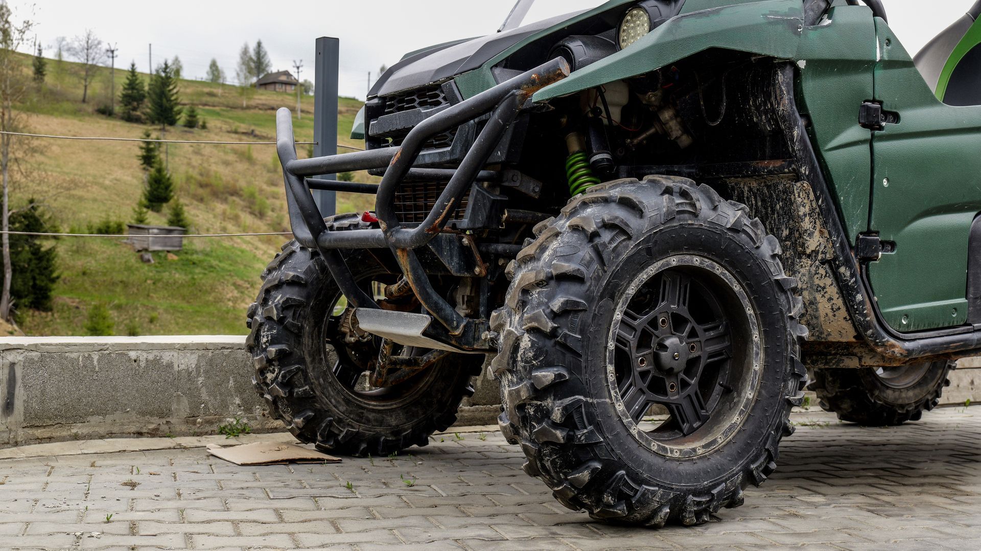 Close-up of a green off-road vehicle with large muddy tires and a rugged front bumper on a paved path outdoors Close-up of a green off-road vehicle with large muddy tires and a rugged front bumper on a paved path outdoors
