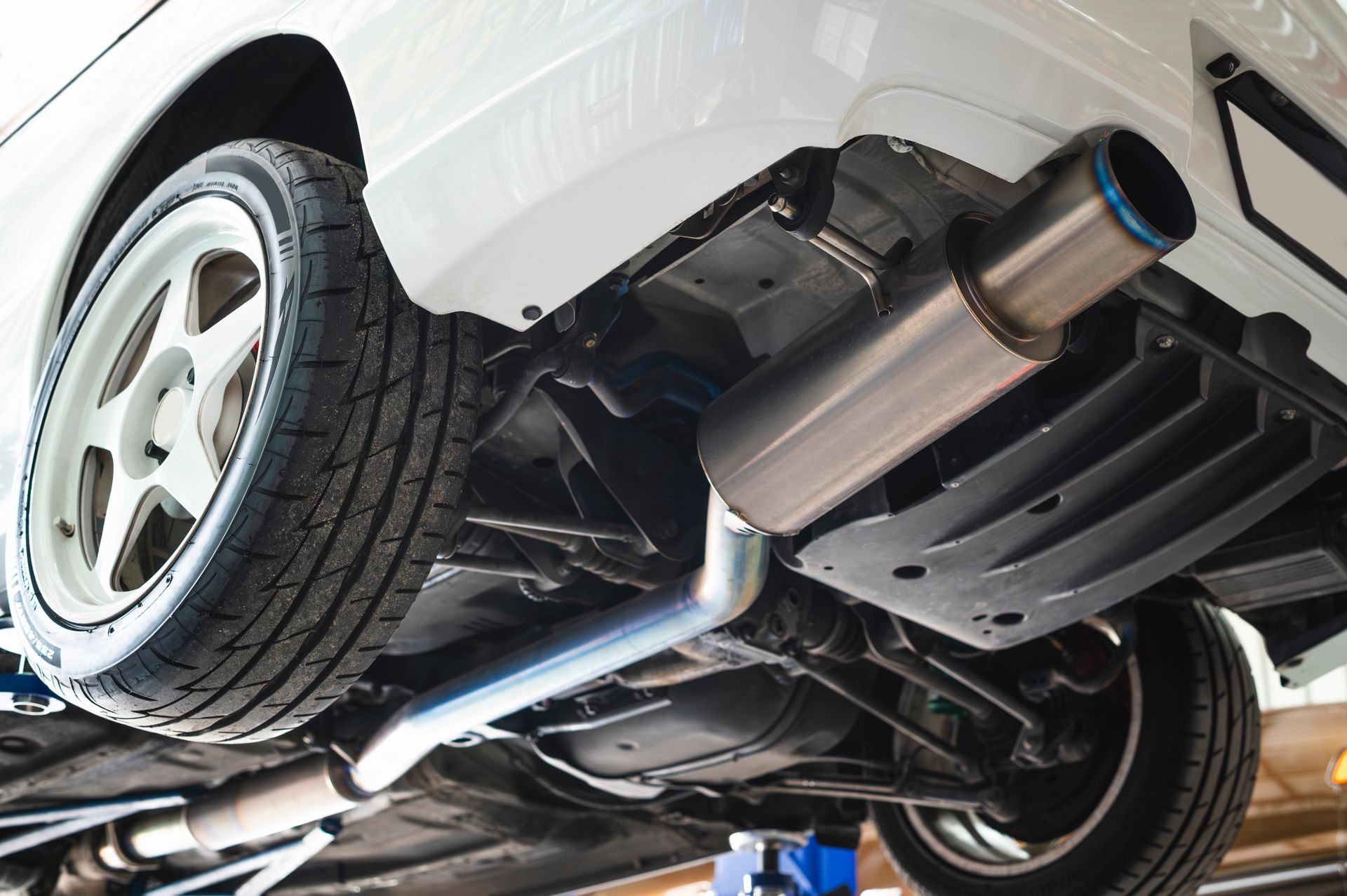 Underside of a white car on a lift, showing the exhaust system and rear wheel in a garage Underside of a white car on a lift, showing the exhaust system and rear wheel in a garage