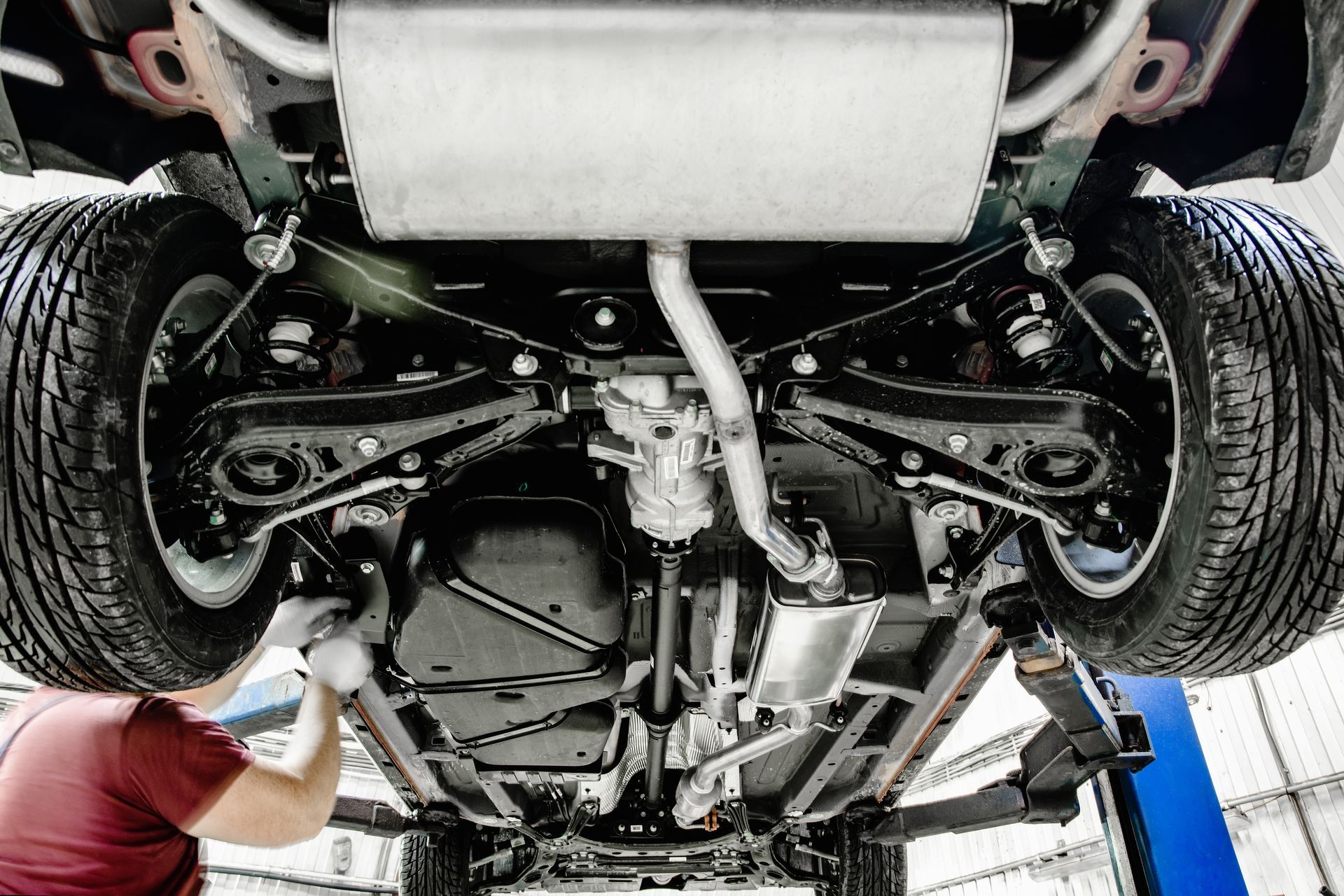 Underside of a car on a lift, with a mechanic inspecting the suspension and exhaust system. Underside of a car on a lift, with a mechanic inspecting the suspension and exhaust system.