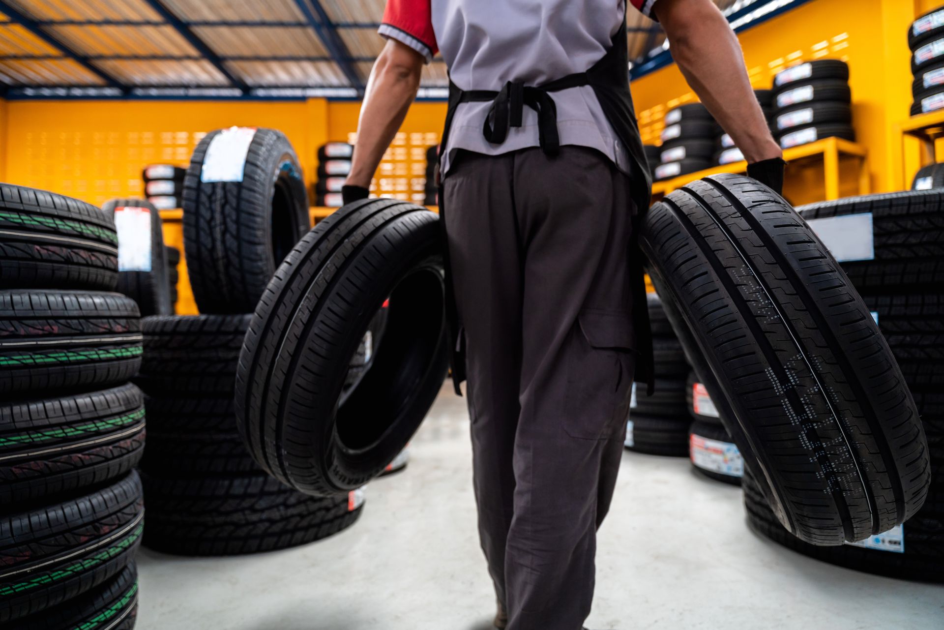 Person carrying two tires in a bright tire shop or warehouse with stacks of tires in the background Person carrying two tires in a bright tire shop or warehouse with stacks of tires in the background