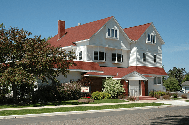 White two-story house with red roof. Front porch, tree, and green grass. Bright sunny day.