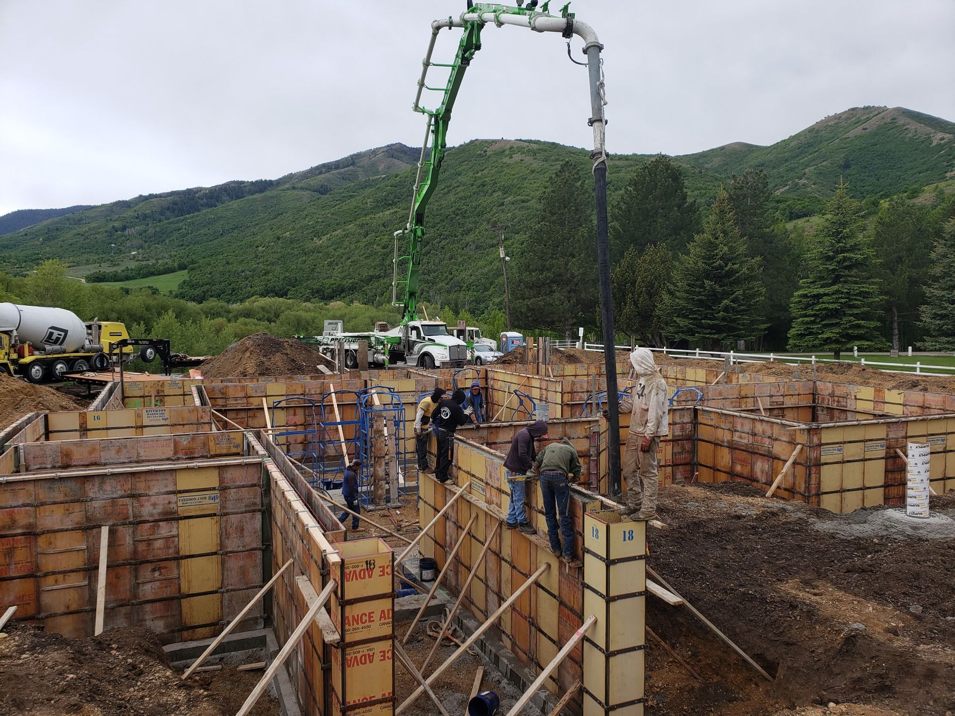 Construction site with workers, rebar forms, and a concrete pump against green hills.