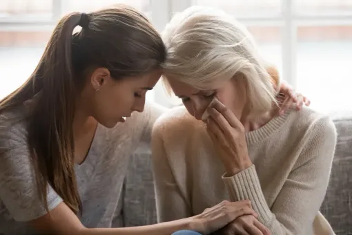 Two women sitting close together, one comforting the other as she wipes away tears.