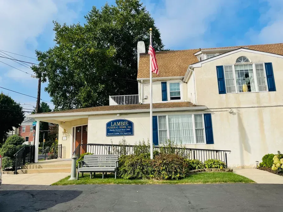 Small beige building with blue shutters, a flagpole, and a blue sign in front under a leafy tree.