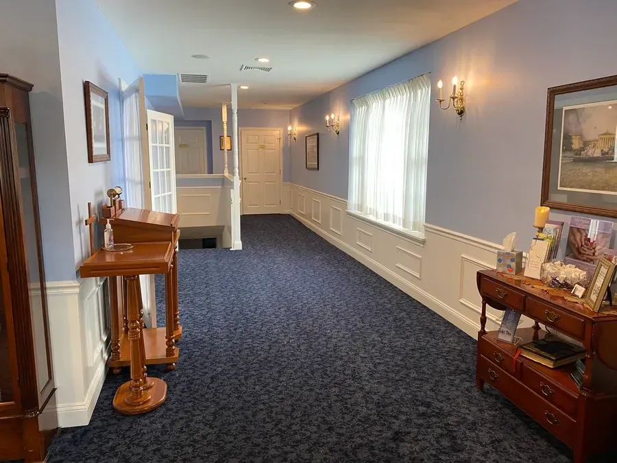 Long carpeted hallway with blue walls, windows, wall sconces, and wooden furniture and paintings.
