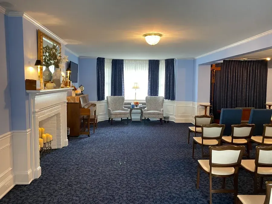 Bright meeting room with blue carpet, white chairs, fireplace, and windows with dark curtains.