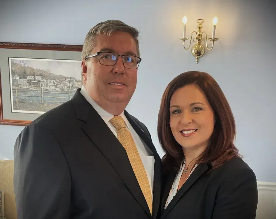 Two smiling colleagues in suits standing indoors by framed art and a wall sconce.