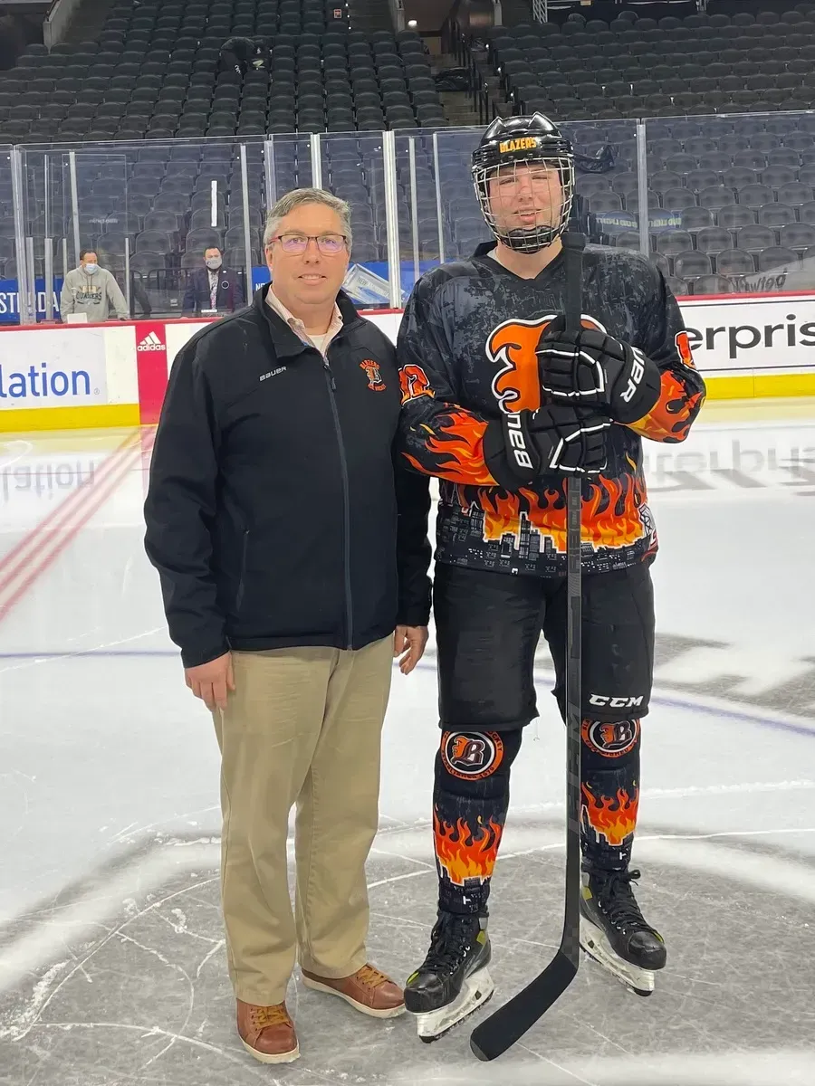 Two hockey players on the ice, one in black and orange gear holding a stick, standing beside another man.