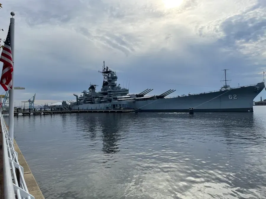 Battleship docked in calm harbor, seen from the waterfront under a cloudy sky