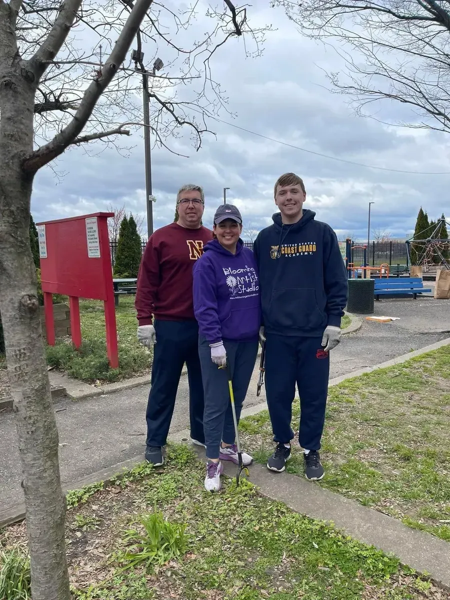 Three people standing outdoors by a tree and red sign, wearing purple and navy sweatshirts.