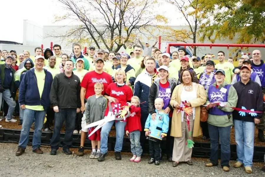 Group of people posing outdoors in front of a red vehicle, many holding shovels at a community event