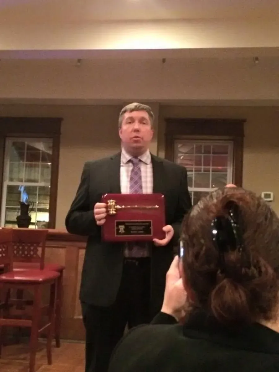 Man in a suit holding a plaque at an indoor event, with an audience member in the foreground