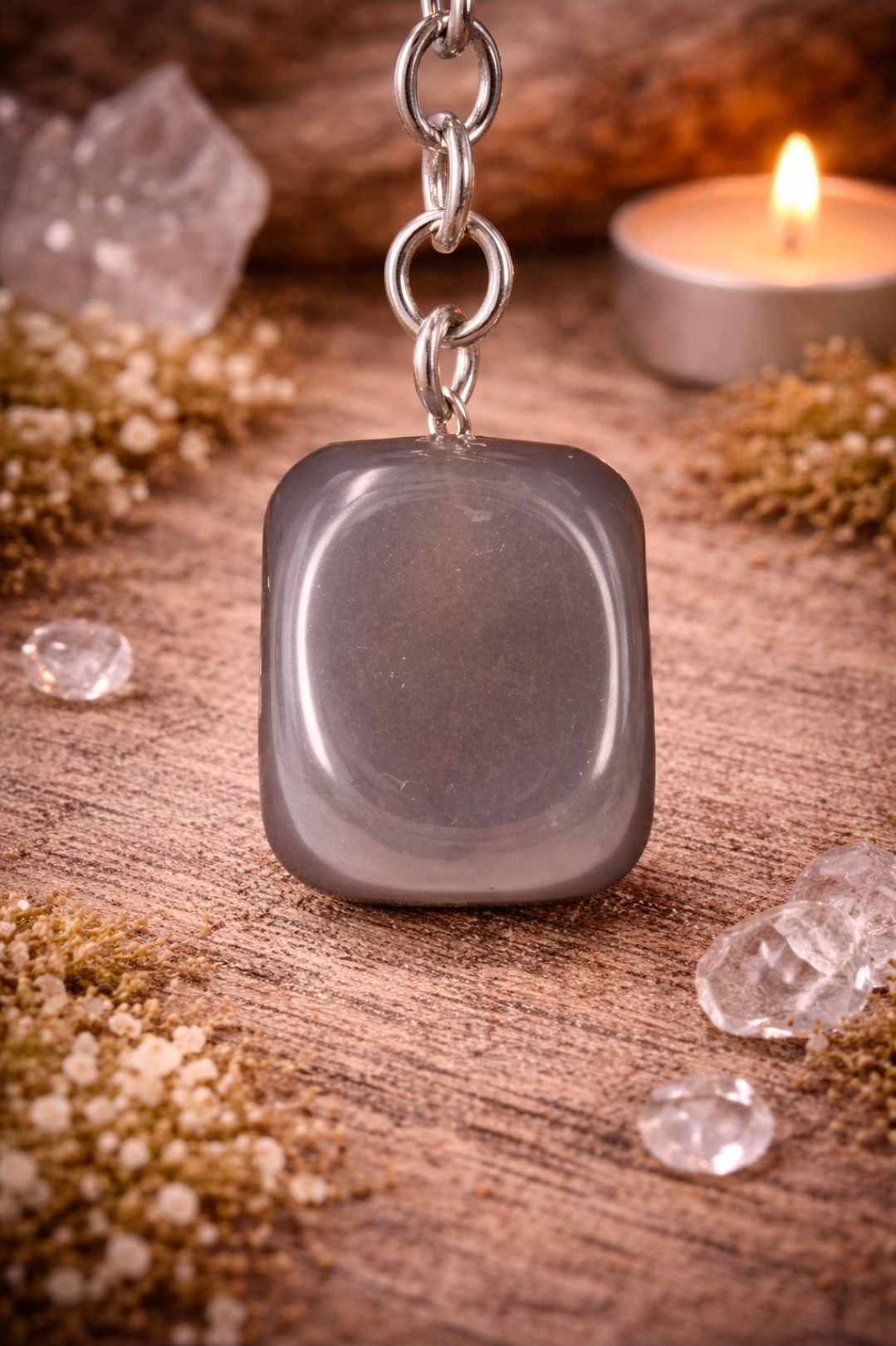Grijze, gepolijste stenen hanger aan een ketting, omringd door kristallen en een kaars op een houten ondergrond.