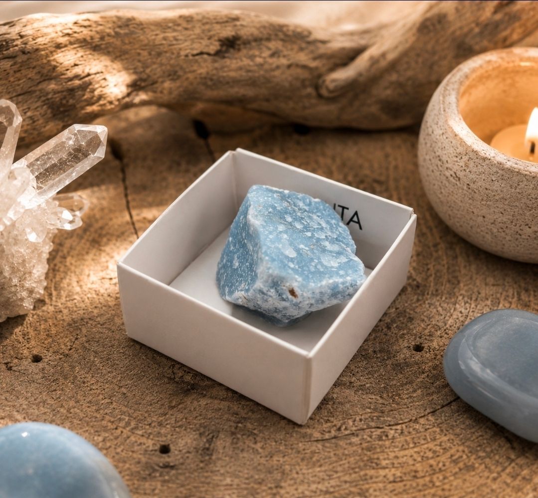 Een blauw kristal in een witte doos op zand, met een kaars, een kwartscluster en een blauwe steen in de buurt.