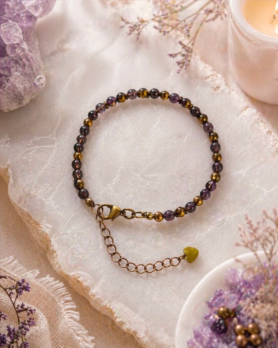 Kralenarmband met paarse en gouden kralen en een bungelende ketting op een zachtroze stoffen achtergrond.