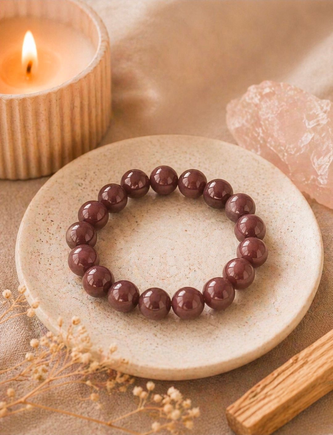 Een bordeauxrode kralenarmband op een beige schaaltje naast een brandende kaars en roze kristallen decoratie.