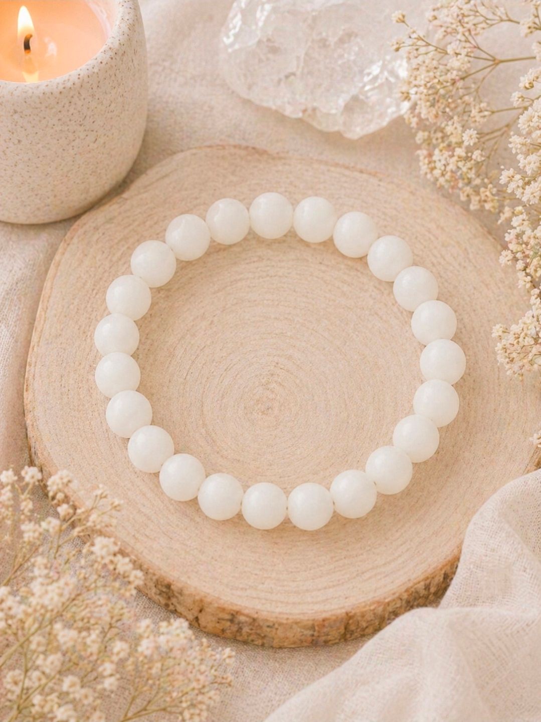 Een witte kralenarmband op een houten onderzetter met zachtbeige decoratie en een brandende kaars.