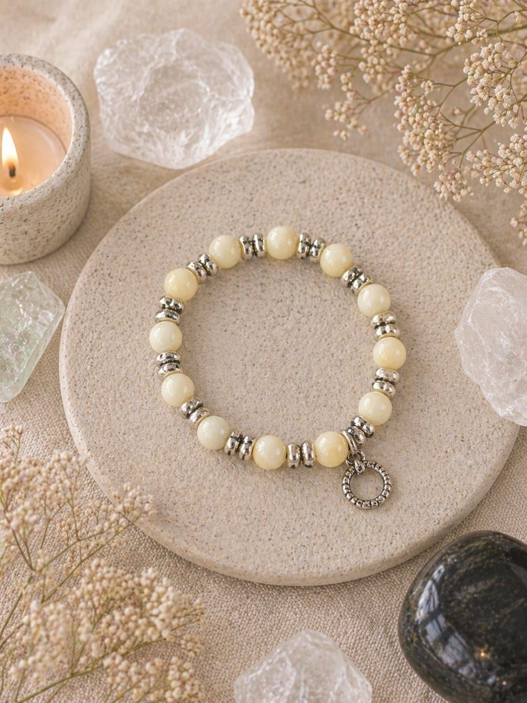 Crèmekleurige kralenarmband met zilveren accenten op een beige stenen dienblad, omringd door kaarsen en kristallen.