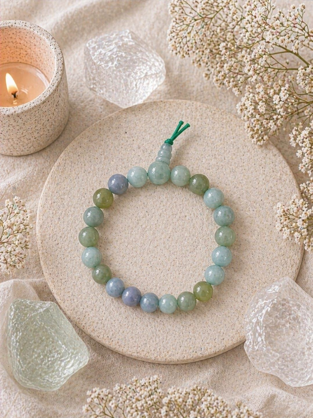 Een groene kralenarmband op een beige bord met een kaars en gedroogde bloemen ernaast.