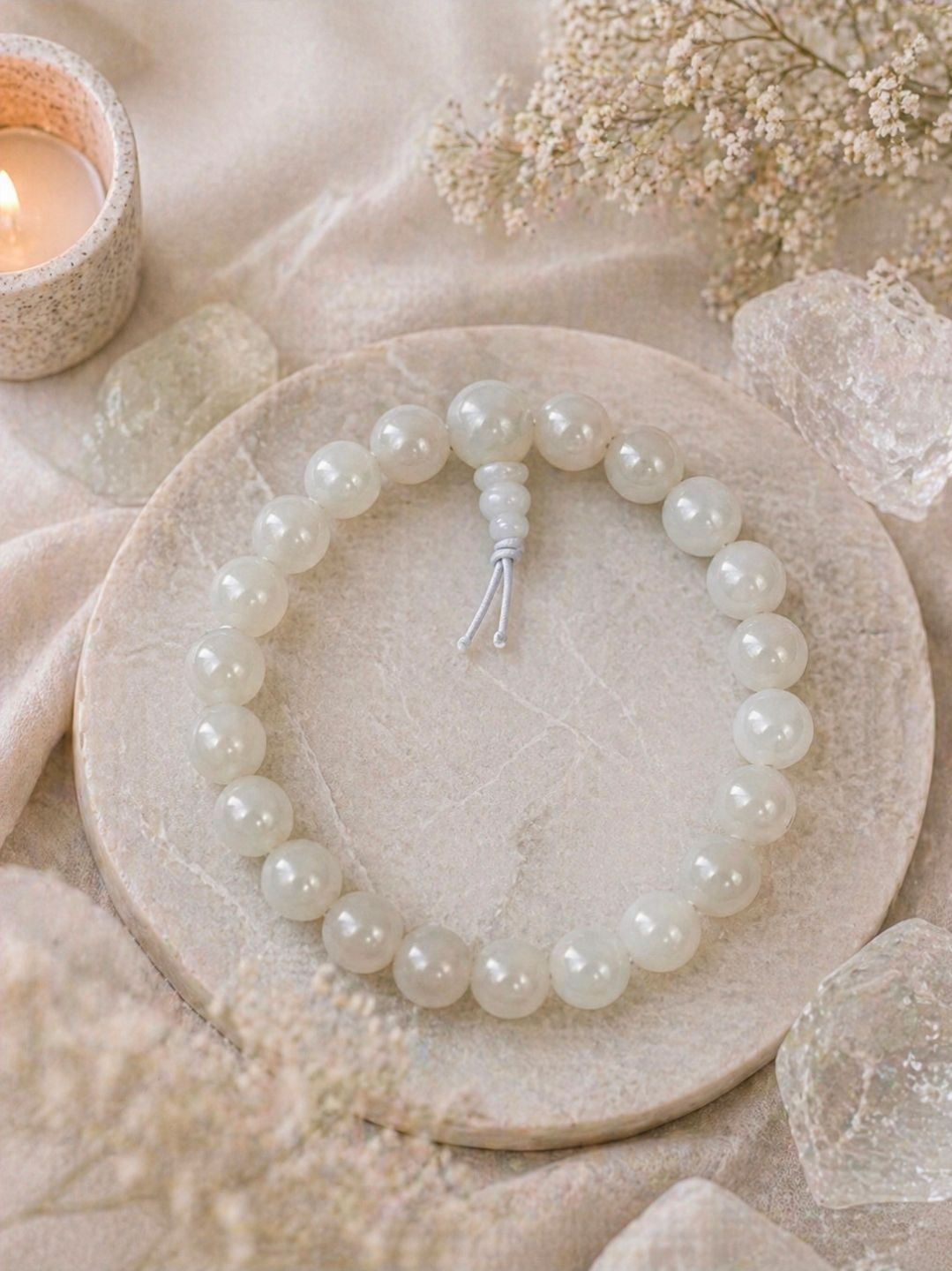 Een witte kralenarmband op een beige bordje naast een brandende kaars en gedroogde bloemen.