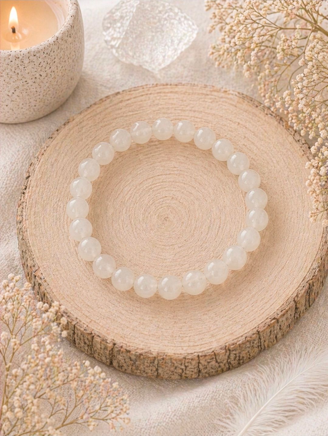 Een witte kralenarmband op een houten schijfje naast een brandende kaars en zacht pampasgras.