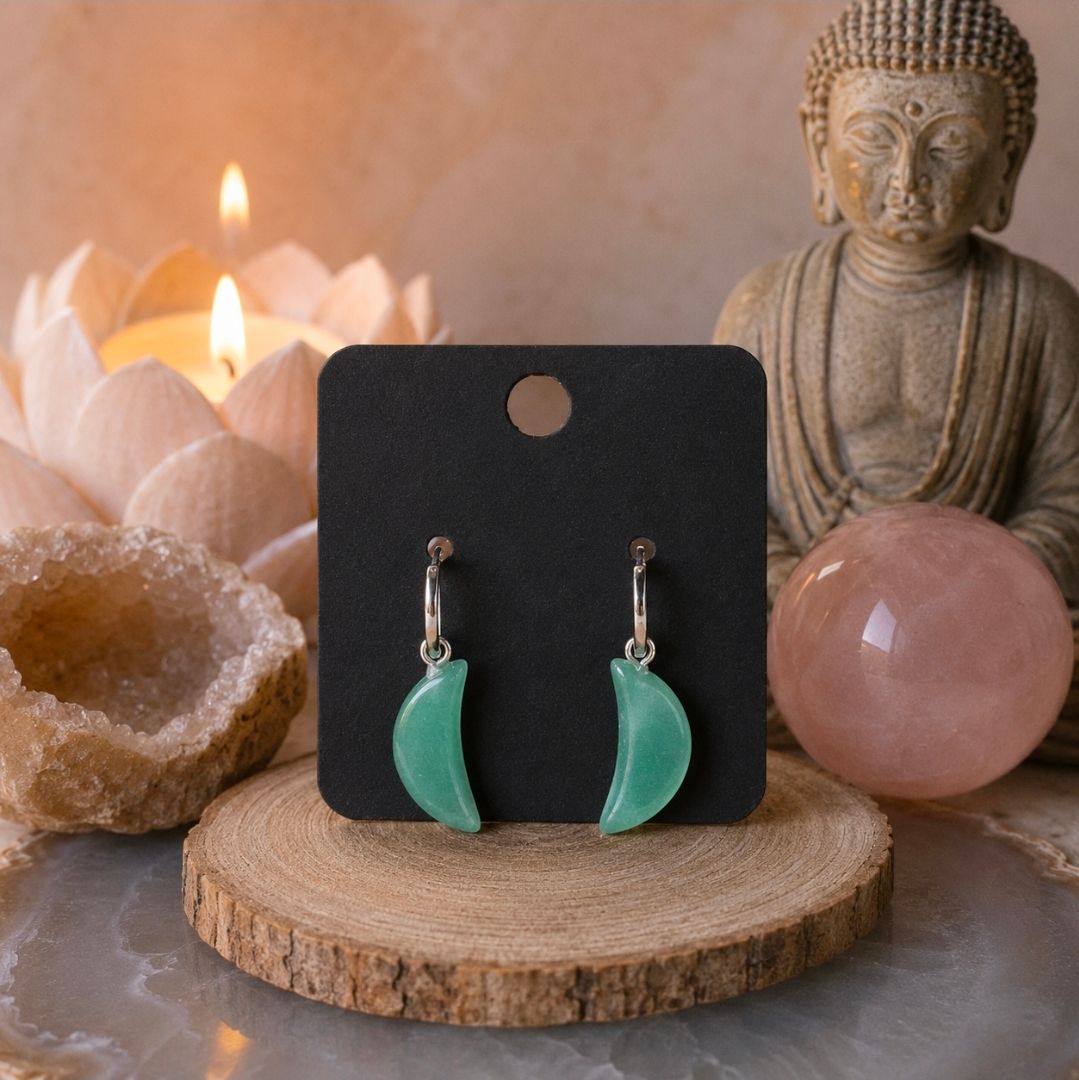 Groene halvemaanvormige oorbellen op een zwart presentatiekaartje, gestyled met kaarsen, kristallen en een Boeddhabeeld.