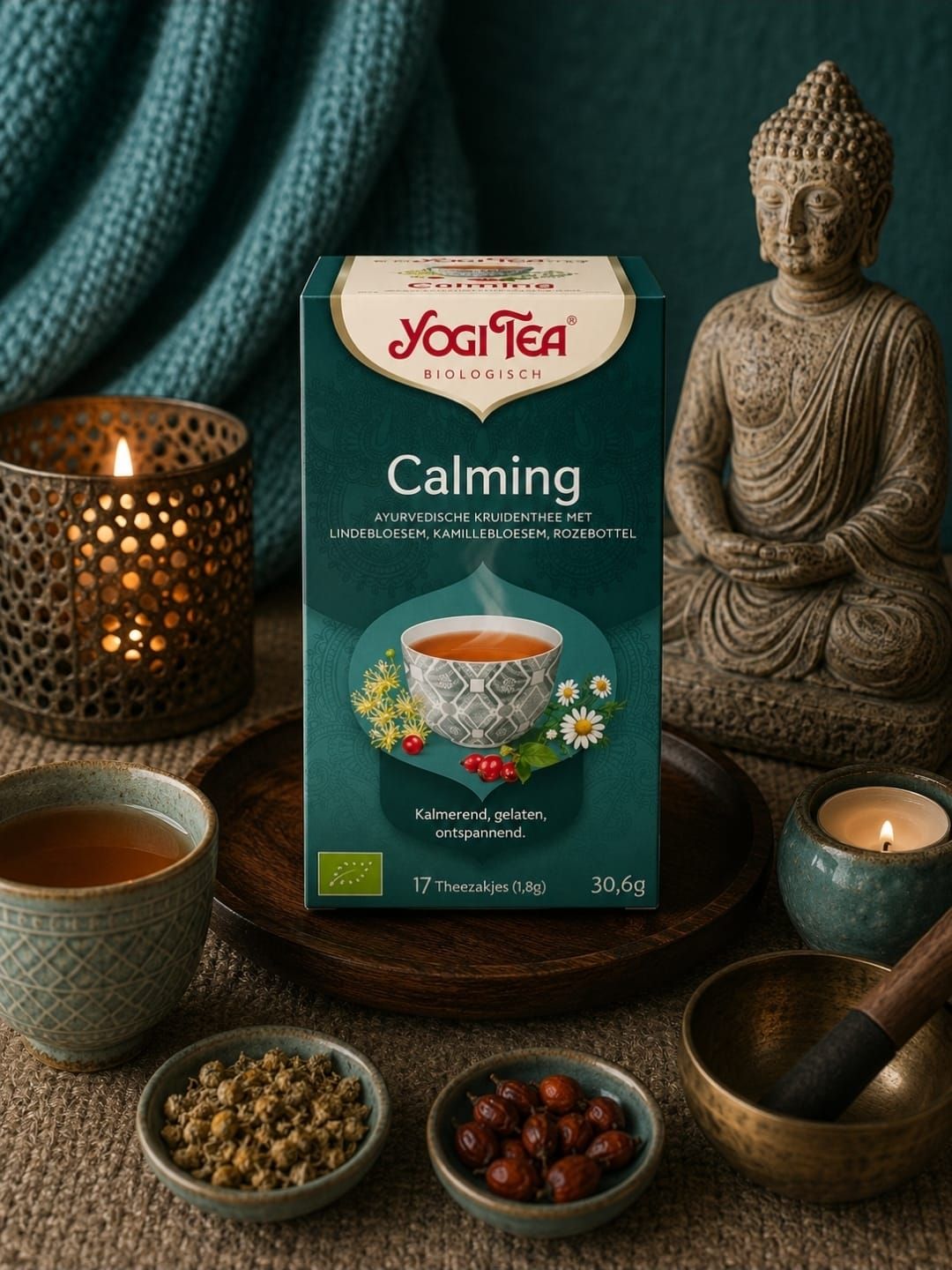 Een Yogi Tea Calming-doosje naast een theekopje, kruiden, een kaars en een klankschaal op een donkerblauwe tafel.