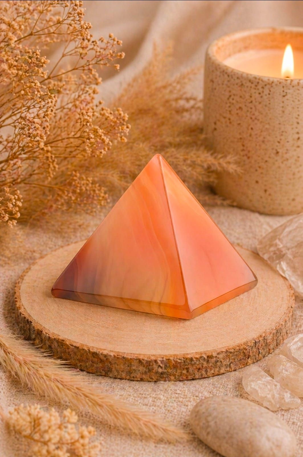 Een roze kristallen piramidekaars op een rond houten schijfje naast een gloeiende kaars in een warme beige omgeving.
