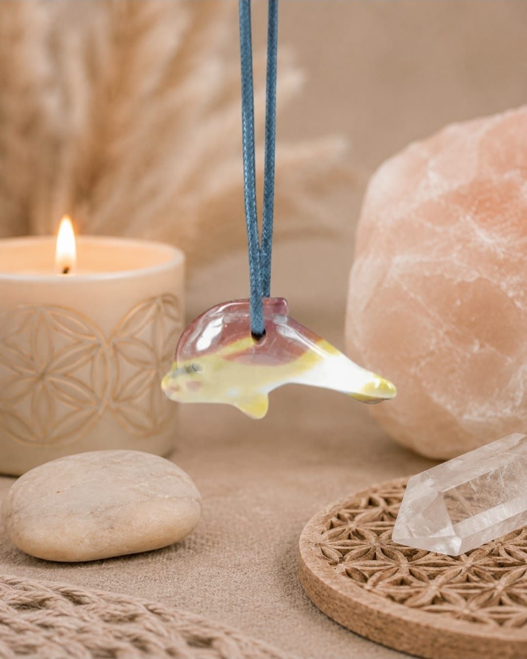 Een ketting met een opaalachtige hanger hangt naast een brandende kaars, kristallen en een geweven dienblad op een beige ondergrond.