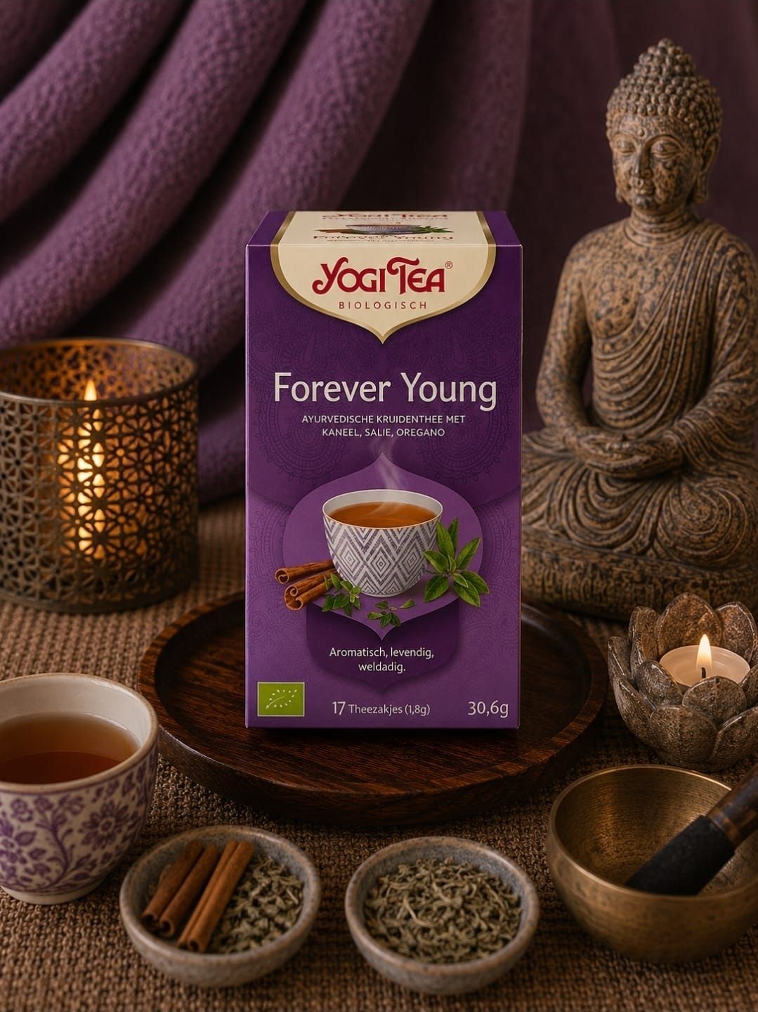 Yoga Tea “Forever Young”-doos met kaars, Boeddhabeeldje en ingrediënten voor kruidenthee op een paarse doek.