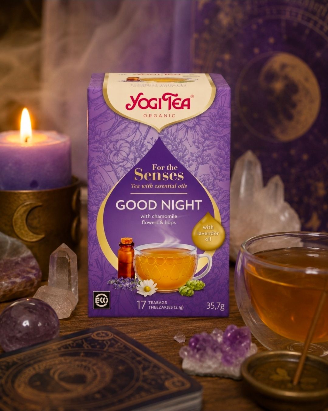 Een doosje Yogi Tea Good Night kruidenthee naast een brandende paarse kaars en een kristal op een donkere, mystieke tafel.