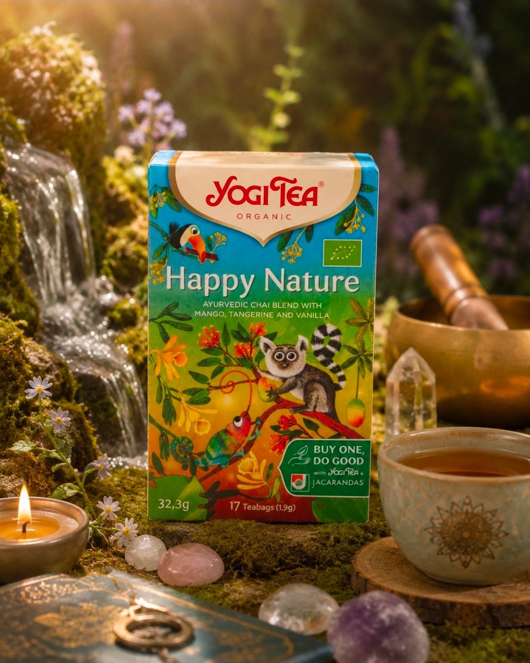 Een doos Yogi Tea Happy Nature naast een theekopje, een kaars en een waterval in een warme, groene omgeving.