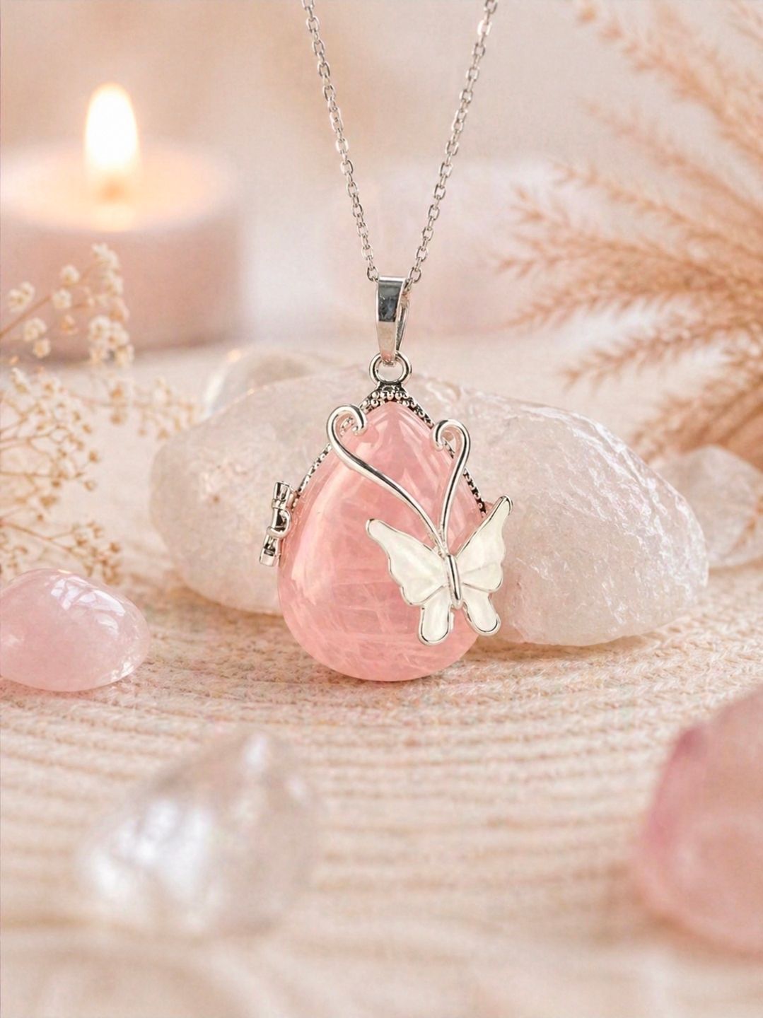 Roze kristallen vlinderhanger aan een ketting op een zacht pastelkleurige stof, met kaarsen en decoratieve bloemen erbij.