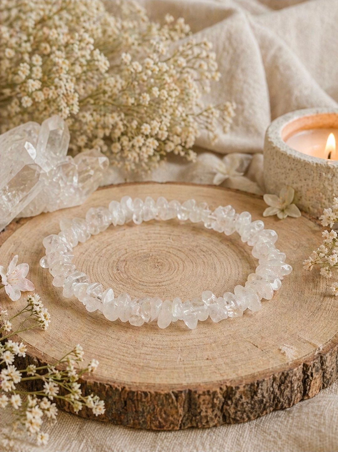 Kristallen kralenketting op een houten schijfje naast gedroogde bloemen en een brandende kaars op een zachtbeige achtergrond.