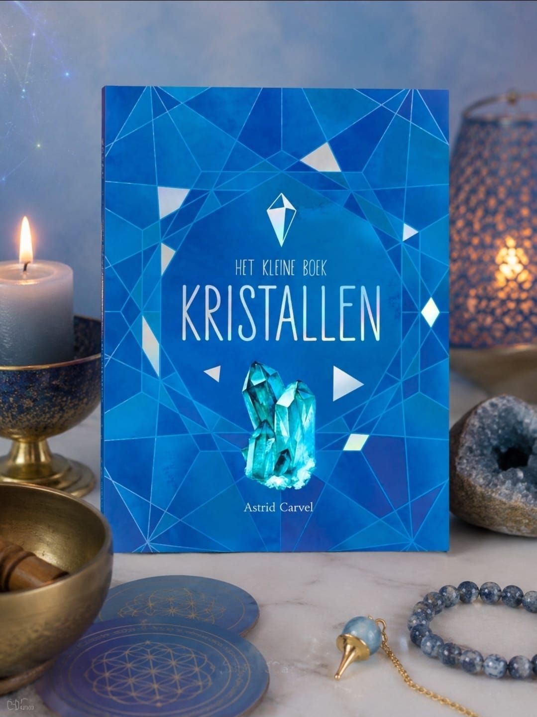 Een boek over blauwe kristallen met een geometrische omslag, tentoongesteld naast kaarsen, kristallen voorwerpen en sieraden