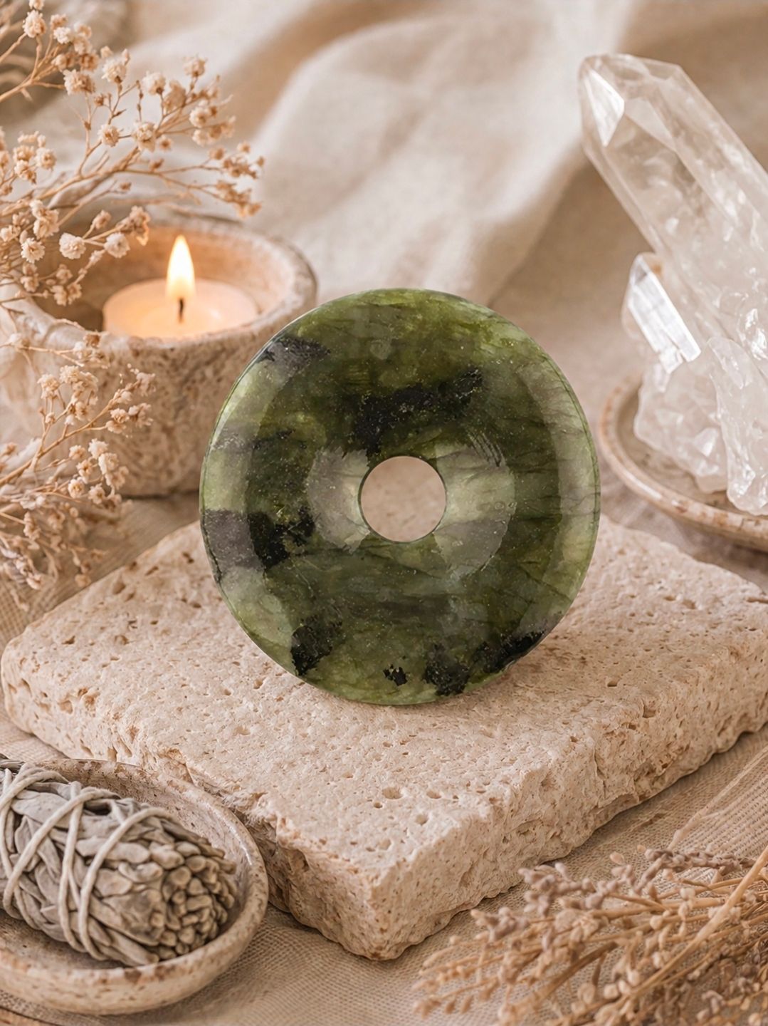 Groene stenen donut op een beige, gestructureerd blok naast een brandende kaars en een helder kwartskristal.