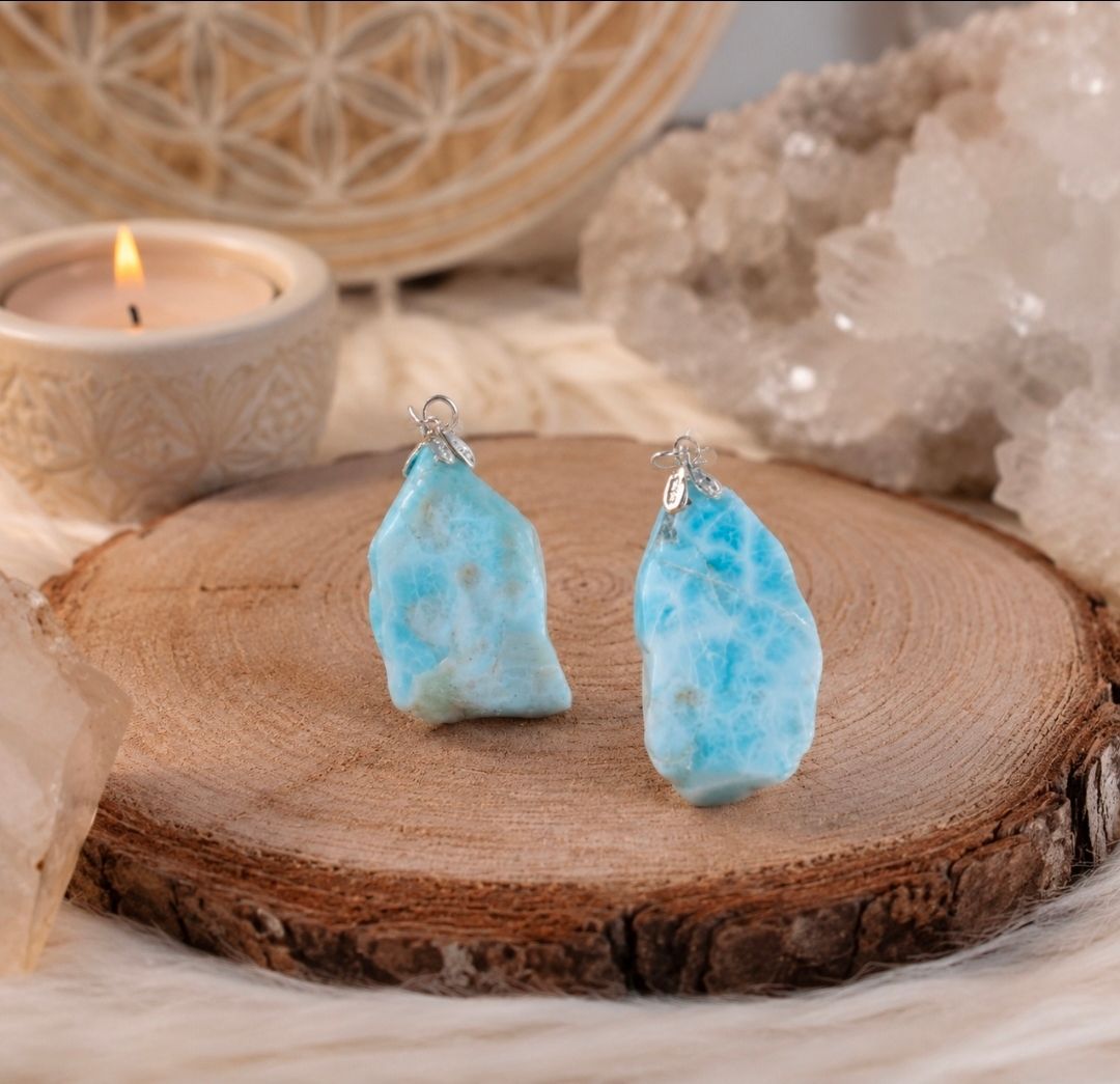 Twee blauwe kristallen hangers op een houten schijf met een brandende kaars en kwartskristallen op de achtergrond.