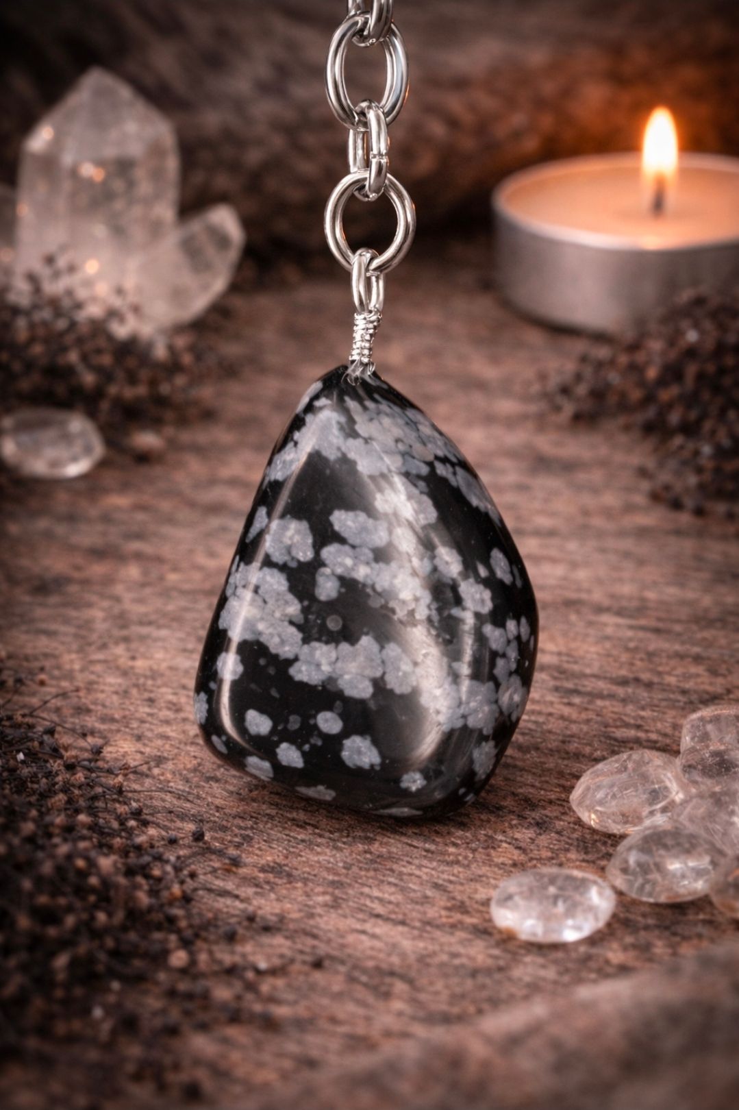 Sneeuwvlokvormige obsidiaan hanger aan een ketting met kaarsen en kristallen op een houten ondergrond.