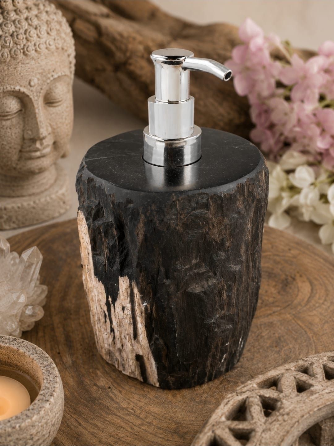 Rustieke zwarte zeepdispenser met zilverkleurige pomp op een houten ondergrond naast een Boeddhabeeld en bloemen.