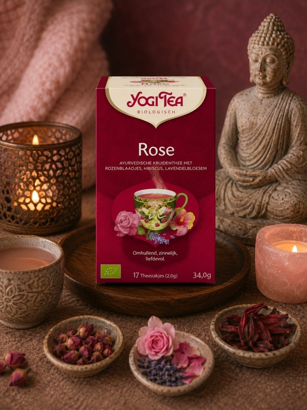 Yogi Tea Rose-doos met rozenblaadjes en theekopjes, kaarsen en een Boeddhabeeld op een roze achtergrond.