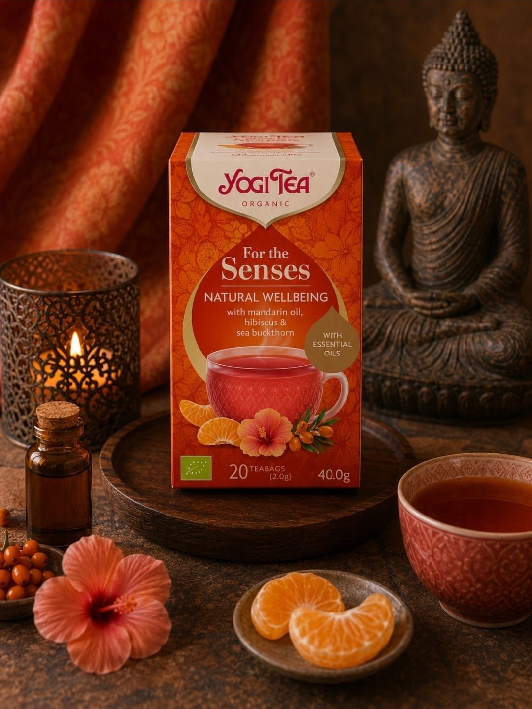 Yogi Tea-box met Boeddhabeeldje, kaars, bloemen en sinaasappelschijfjes in een warme, spa-achtige setting.